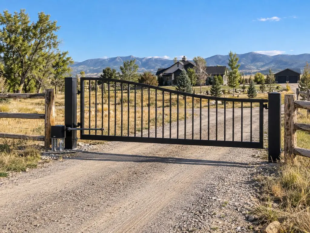 Black single swing gate with a post-mounted arm operator across a gravel drive, split-rail fence either side